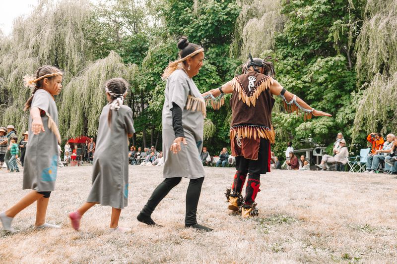 Coast Salish dancers including young girls and a feathered regalia performer move across the grass