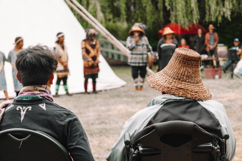 Two community members watch the ceremony, one wearing a woven cedar hat