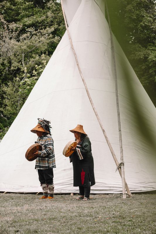 A line of young dancers in grey ceremonial dresses stand before the tipi