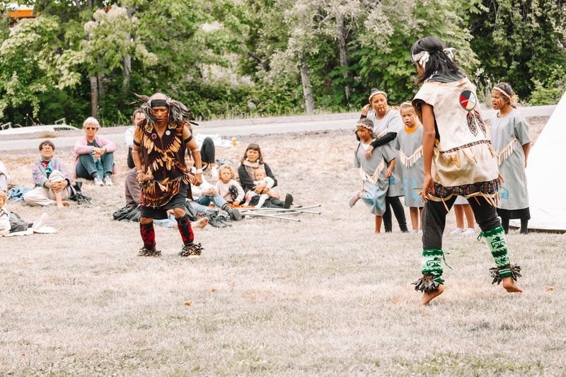 Two community members in cedar hats holding hand drums in front of the tipi