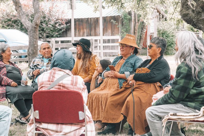 A community member with a feather and woven cedar hat watches from a green camp chair