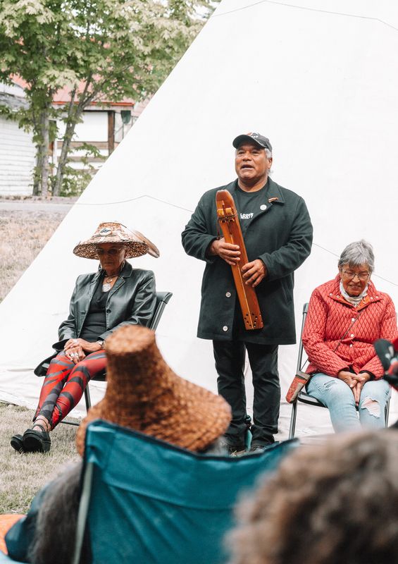 A Knowledge Keeper standing and speaking with hand on heart, an Elder seated beside