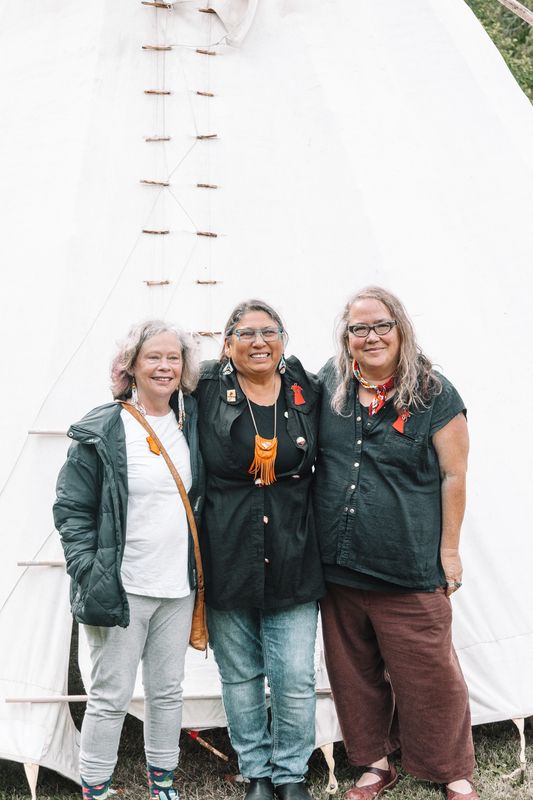Three women smiling and standing together in front of the tipi