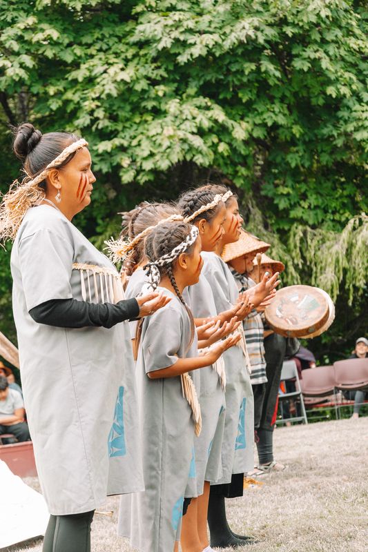 A line of mothers and daughters in grey regalia dancing together near the tipi