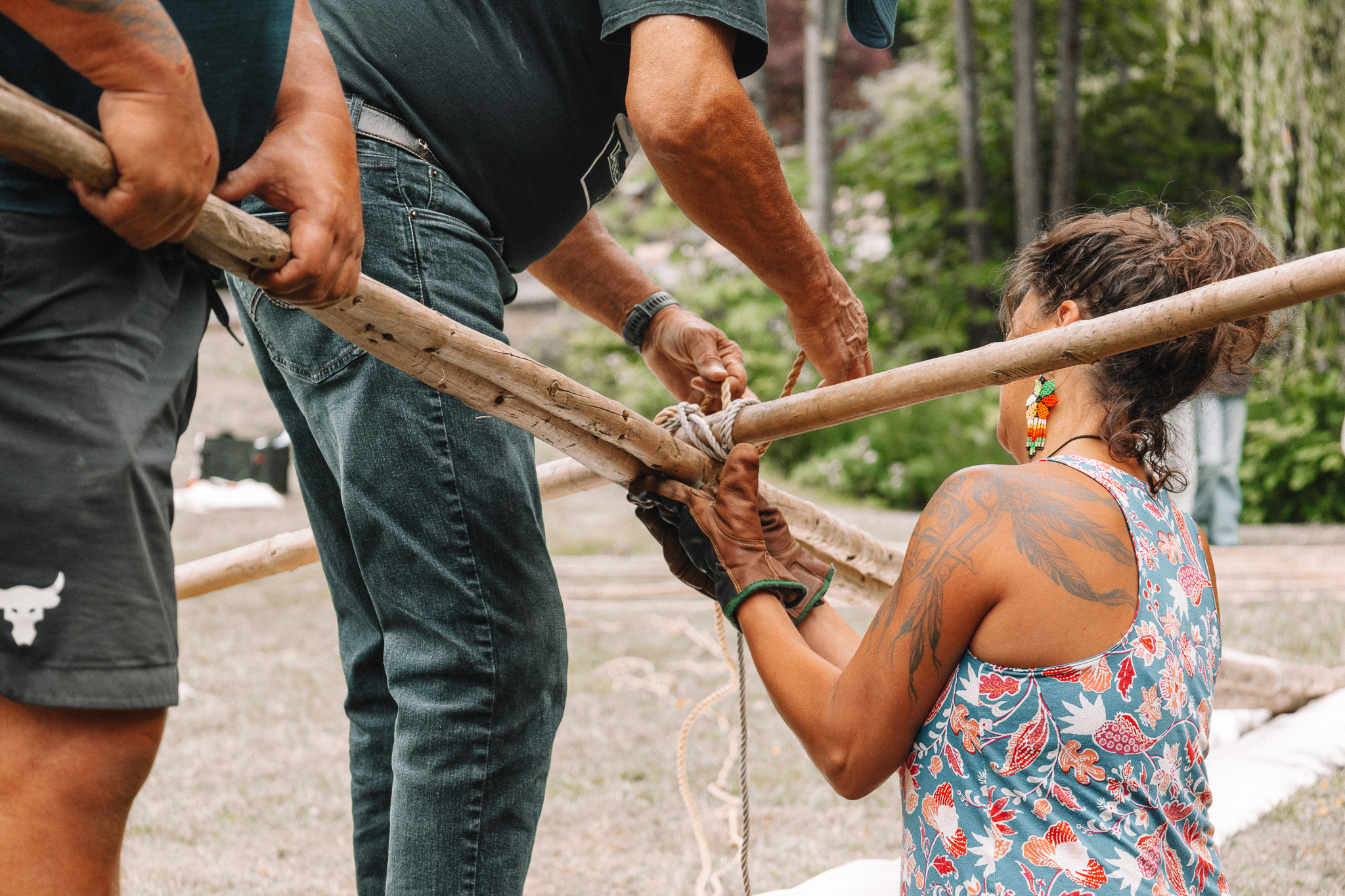 Community hands tying lodgepoles together with rope at the tipi raising — a young woman in a floral tank with beaded earrings and dragonfly tattoos works alongside an Elder
