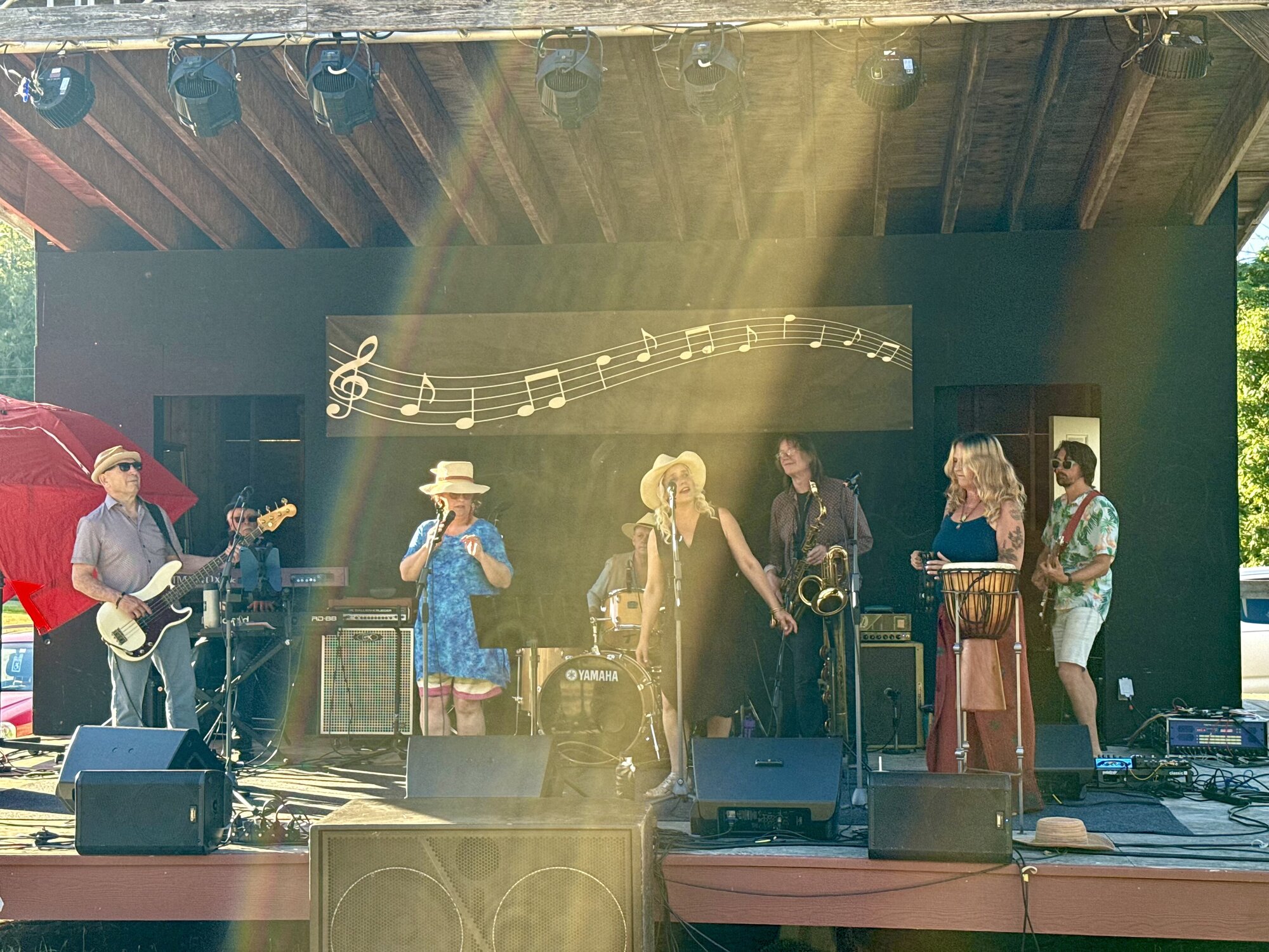 The band Local Motion performing on an outdoor stage in a beam of sunlight, with a flowing music-note backdrop behind them