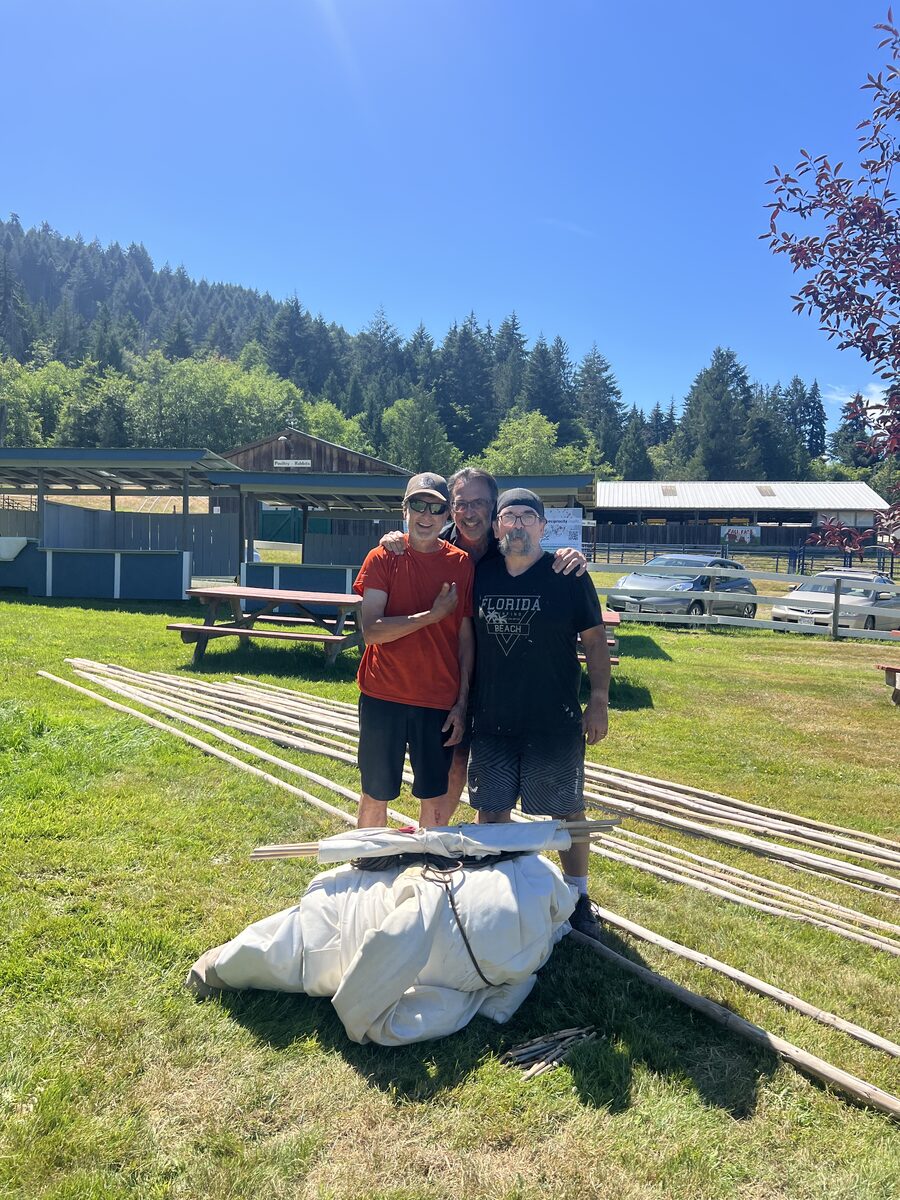 Three men standing together beside the folded tipi cover and poles on the grass at the Farmers' Institute