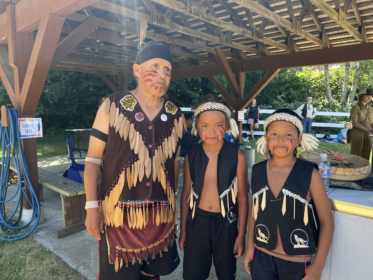 An Elder in feathered regalia with two young dancers in vests and woven headbands at the gathering